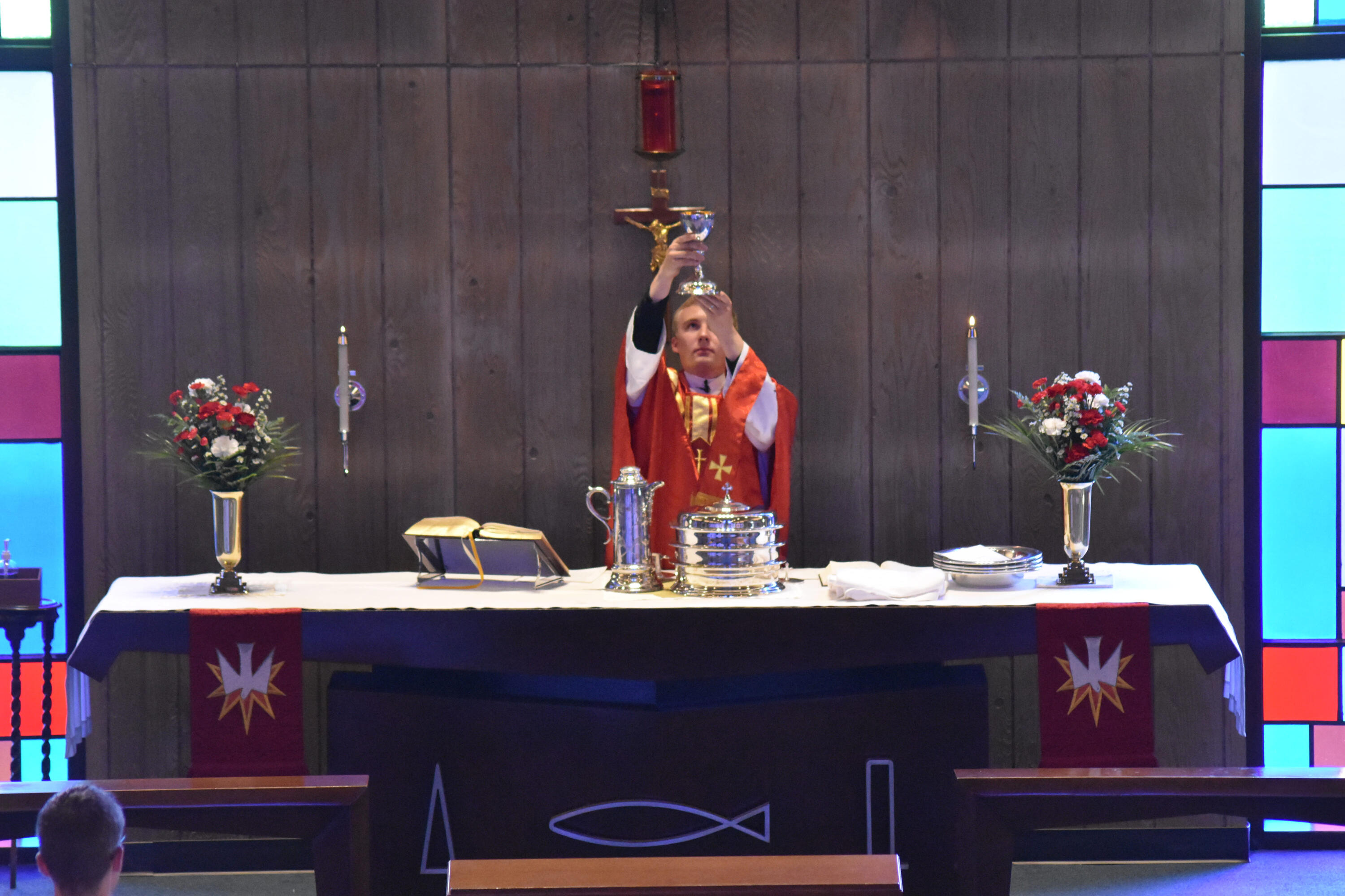 Pastor Otten of Holy Cross in Racine, Wisconsin celebrates communion. Pastor Luke Otten elevates the chalice in a service at Holy Cross Lutheran Church in Racine, Wisconsin. He is wearing a red chasuble. The altar also has red paraments and flowers.