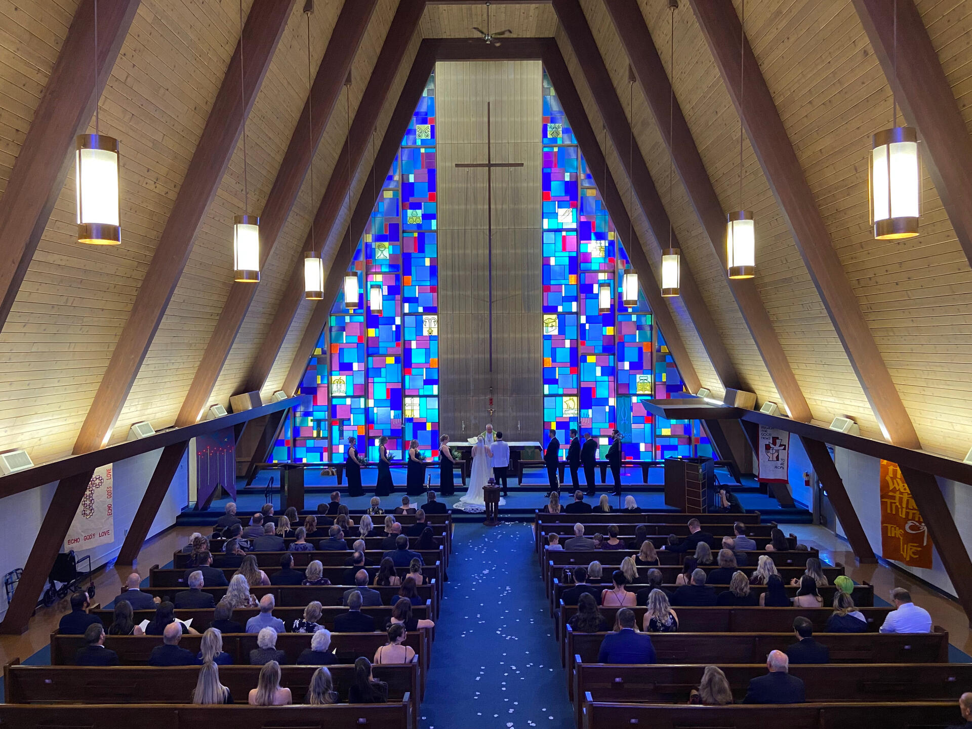 A wedding at Holy Cross Lutheran Church in Racine, Wisconsin. A bride and groom stand in front of the altar at Holy Cross Lutheran Church in Racine, Wisconsin, surrounded by bridesmaids and groomsmen. The blue aisle carpet is covered in flower petals, and the church is full of guests. The blue and purple stained glas